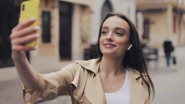 Šťastná hezká dívka s bezdrátovými sluchátky, jak se video hovor drží její smartphone vertikální mávání Hi s úsměvem ukazující palec nahoru. — Stock video