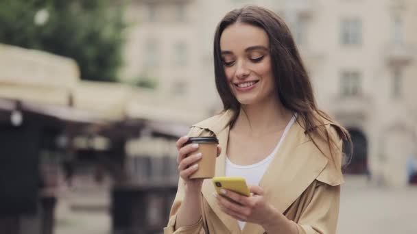 Attraktives junges Mädchen im Trenchcoat mit ihrem Smartphone und lächelnd mit einem Pappbecher Kaffee trinkend vor Altstadthintergrund aus nächster Nähe . — Stockvideo