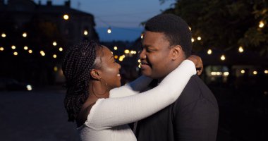 Genç çift sarılıyor ve kameraya bakıyor. Gece caddesinde birlikte duran gülümseyen sevgili ve kız arkadaşa yakın durun. Ligts arkaplanı. Aşk ve ilişkiler kavramı.