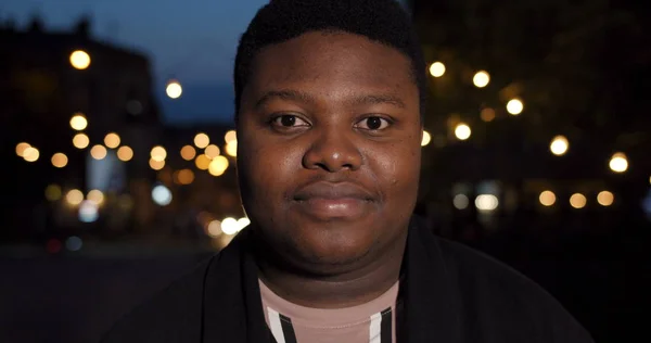 Närbild av fett afrikansk man tittar rakt mot kameran. Ung kille står utomhus. Nattstadens ljus bakgrund. Porträtt. Begreppet människor och nattliv. — Stockfoto