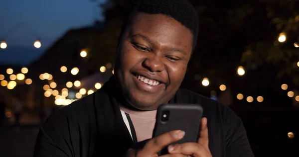 Primer plano de tipo feliz utilizando el teléfono inteligente y de pie al aire libre. Hombre africano charlando en las redes sociales y mirando la pantalla del teléfono. Fondo de luces de ciudad. Concepto de comunicación moderna . — Foto de Stock