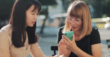 Dışarıda oturan iki kız akıllı telefon ses tanıma sistemi kullanıyor. Neşeli kız, akıllı telefon ses tercümanı uygulaması aracılığıyla yabancı bir arkadaşla konuşuyor. İletişim ve teknoloji kavramı.
