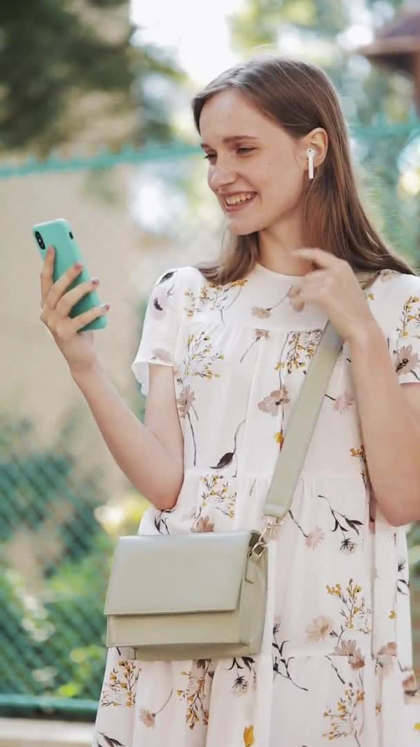 Junge schöne kaukasische Frau in weißem Blümchenkleid mit Kopfhörern, die ihr Smartphone benutzt, um einen Videochat zu führen, in dem sie lächelnd auf der Straße herumwinkt. vertikales Video. — Stockvideo