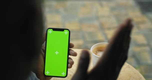 Lviv, Ukraine - Le 09 août 2019 : Gros plan d'un Afro-Américain tenant son Smartphone avec écran vert ayant un appel vidéo avec une tasse de café en papier sur la table en arrière-plan. Vue de l'arrière . — Video