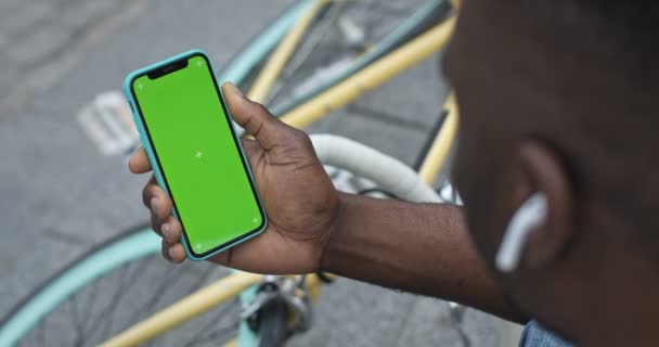 Közelkép afro-amerikai férfi fülhallgató fogja a Smartphone Looking to Green Screen. Ő ül a lépcsőn a városban, stílusos színes kerékpár a háttérben. — Stock videók