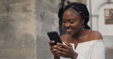 Güzel kız akıllı telefon ekranına bakıyor, şaşırmış yüz ifadeleri takınıyor ve gülümsüyor. Genç ve çekici bir kadın duygularını belli ediyor ve dışarıda dururken akıllı telefon kullanıyor. Eski binalar.