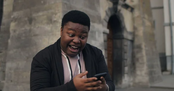 Giovane ragazzo africano facendo faccia sorprendente e guardando lo schermo dello smartphone mentre in piedi all'aperto. Millennial maschio sembra estremamente felice e soddisfatto di buone notizie. Vecchio sfondo della città . — Foto Stock
