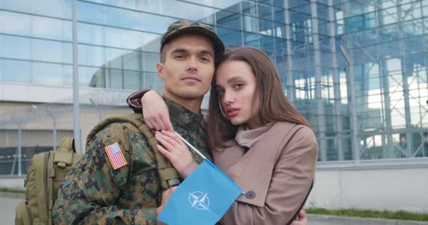 Lviv, Ucrania - 30 de octubre de 2019: Retrato de una pareja con bandera de la OTAN mirando a la cámara. Primer plano de la joven y su novio militar de pie cerca del aeropuerto. Retratos de personas . — Vídeos de Stock