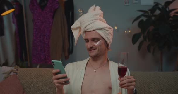 Close up of young transgender in rob and towel on head using phone and making jealous face. Man trans drinking wine while sitting on couch and surfing net. Home background. — 비디오