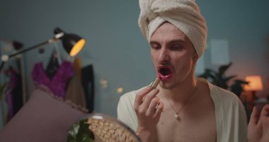 Erkek travesti diva otururken, pembe ruj sürerken ve gösteriye hazırlanırken. Genç adam travesti ve aynaya bakarken makyaj yapıyor ve başını hareket ettiriyor..