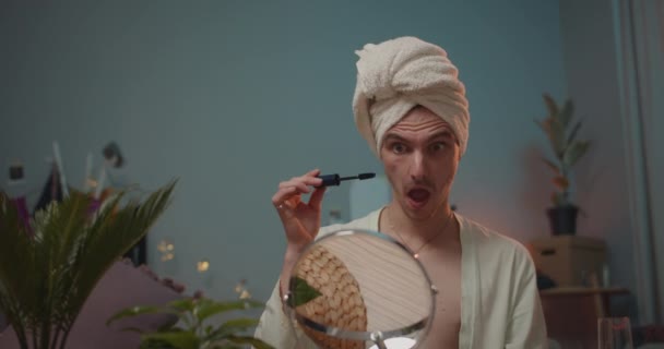 Νέοι τρανσέξουαλ εφαρμογή μάσκαρα στις βλεφαρίδες με ανοιχτό στόμα. Άντρας trans με ρόμπα μπάνιου κάνει μακιγιάζ και κοιτάζοντας στον καθρέφτη, ενώ ετοιμάζεται για το κόμμα. Ιστορικό δωματίου. — Αρχείο Βίντεο