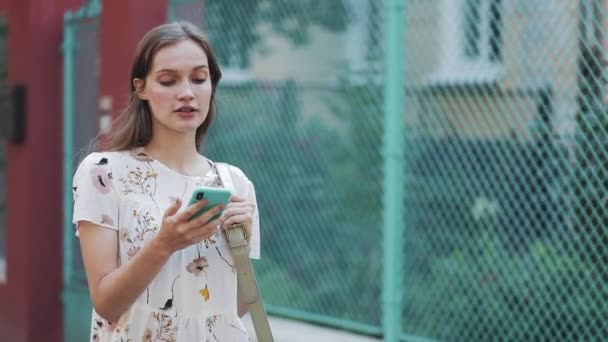 Bastante joven caucásica Brown Hair Girl usando vestido blanco de la flor con el bolso en su hombro usando la celebración de su teléfono inteligente mirando sorprendido caminando cerca de la cerca de la turquesa sonriendo . — Vídeo de stock