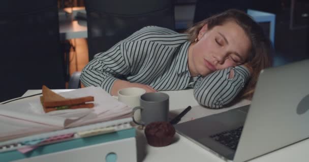 Mujer de negocios agotada tumbada en el escritorio con el ordenador portátil abierto delante de ella. Chica cansada que se queda dormida mientras trabaja largas horas en la oficina nocturna vacía. Concepto de exceso de trabajo . — Vídeos de Stock