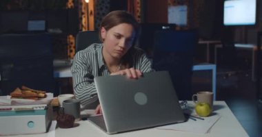 Yorgun kadın ofis çalışanı dizüstü bilgisayarını kapatıp kollarını birbirine vuruyor. Stresli iş kadını, gece geç saatlere kadar boş ofiste çalışırken yaşadığı zorluklardan dolayı bitkin görünüyor.