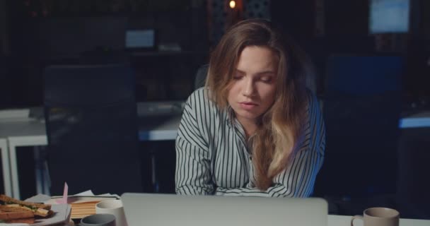 피곤 한 아름다운 여자가 밤늦게 까지 사무실에 있는 동안 카메라를 보고 있는 모습. 예쁜 여성 사무실 매니저가 컴퓨터를 사용하고 있습니다. 직업 과 일에 대한 개념. 사무실 배경. — 비디오