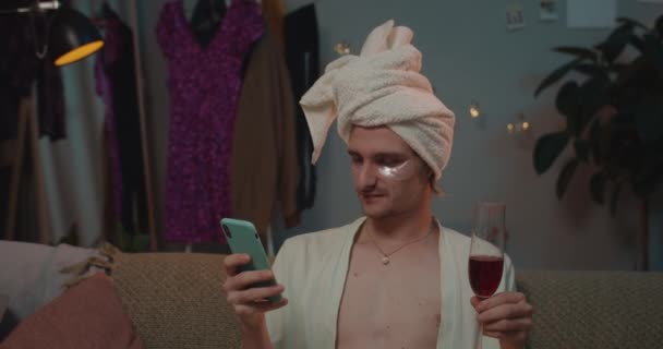Κοντινό πλάνο των χαλαρών trans άνθρωπος στο σπίτι φορούν χρησιμοποιώντας smartphone και πίνοντας κρασί. Νέοι τρανσέξουαλ απολαμβάνουν ελεύθερο χρόνο, ενώ κάθεται στον καναπέ και surfing net. Αρχική φόντο. — Αρχείο Βίντεο