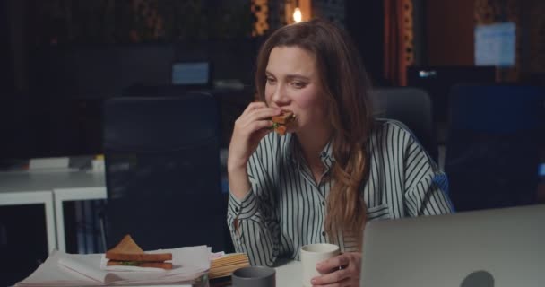 Frustrato donna ufficio manager mangiare panino e bere caffè presso l'ufficio notturno. Ragazza esausta avendo pasto tardi mentre si lavora lunghe ore in worplace vuoto. Concetto di lavoro eccessivo . — Video Stock