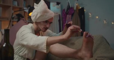 Kanepede otururken ayak tırnaklarına oje süren konsantre bir transseksüel erkek yakın çekim. Genç trans adam güzellik prosedürleri yapıyor ve partiye hazırlanırken şarap içiyor. Oda arkaplanı.