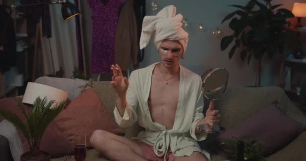 Młody transseksualista w dobrym nastroju pijący wino i palący papierosa patrząc w lustro. Trans-man w szlafroku i ręczniku na głowie siedzi na kanapie i śpiewa piosenkę. — Wideo stockowe