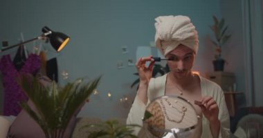 Banyo cüppeli genç travesti randevuya hazırlanıyor ve makyaj yapıyor. Kirpik olarak rimel kullanan ve küçük aynaya bakarken çikolata yiyen bir transseksüel..