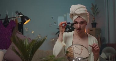 Adam kirpiklere rimel sürüp çikolata yiyor. Adam banyo cüppeli transseksüel makyaj yapıyor ve partiye hazırlanırken aynaya bakıyor. Oda arkaplanı.
