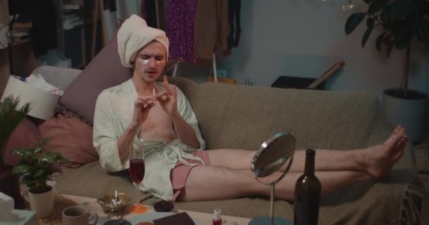 Transgender v saténovém županu relaxovat na pohovce a dělat kosmetické procedury při poslechu hudby.Mladý muž transgender ležící na gauči pilou nehty a zpěv v pilníku nehtů. — Stock video