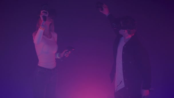 Joven y mujeres usando gafas de realidad virtual sosteniendo joysticks Controladores, moviendo las manos, merodeando de pie en la habitación con fondo de colores de iluminación de neón . — Vídeo de stock