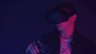 Close Up of Attractive Man in Virtual Reality Glasses Draws Abstract Lines and Figures Using Controllers, Standing in the Room with Neon Lighting Colors Futurictic Background.