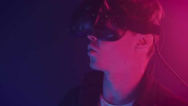 Close Up Portrait of Impressed Guy Wearing Virtual Reality Glasses Looking Up and Smiling, Saying WoW Standing in the Room with Abstract Neon Lighting Colors Background. — Wideo stockowe