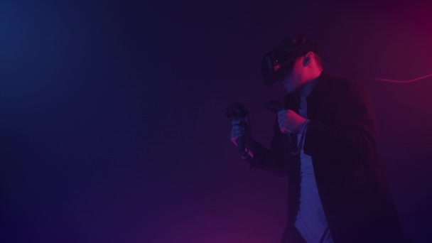 Молодой человек носит гарнитуру виртуальной реальности, коробки, движется руки, тренировки ногами, холдинг джойстики контроллеры стоят на неон освещения цвета футуристического фона . — стоковое видео