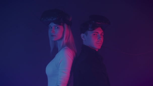 Close Up Retrato de dois jogadores Jovem e garota em pé no fundo de iluminação de néon cibernético com óculos de realidade virtual em sua cabeça, Segurando controladores, Olhando para a câmera . — Vídeo de Stock