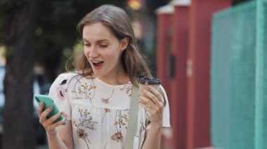 Beautiful Young Woman Wearing White Flower Dress with Bag Cross her Shoulder Using Holding Her Smartphone and Coffee Cup Looking Amazed and Happy Walking near Turquoise Fence Smiling.