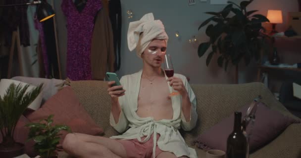 Man transgender in loungewear en undereye pleisters zittend op de bank en wijn drinkend. Jonge transgender ontspannen thuis tijdens het gebruik van de telefoon, op zoek naar camera en glimlachen. — Stockvideo