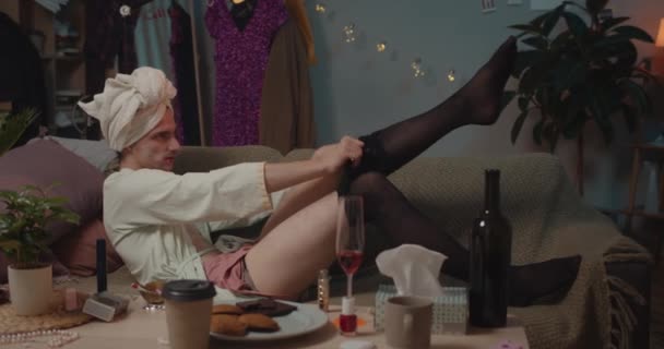 Mężczyzna transgenderowy szykuje się na randkę leżąc na sofie i próbując założyć czarne rajstopy. Domowe tło. Pojęcie ludzi, piękna i czasu wolnego. — Wideo stockowe