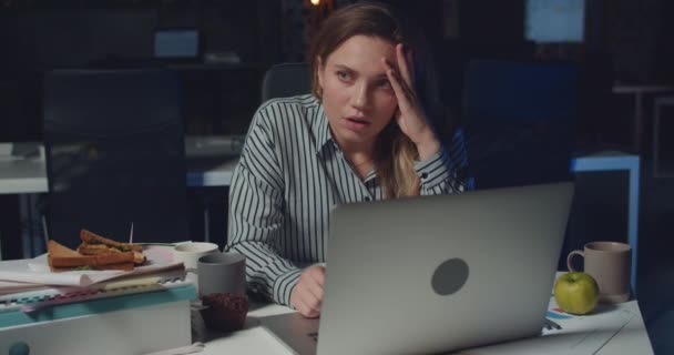 Вид спереду втомленої перевтоми жінки, яка сидить і використовує ноутбук наприкінці офісу. Красива дівчина відчуває себе виснаженою і потирає голову, перебуваючи на самоті на робочому місці. Концепція перевантаження . — стокове відео