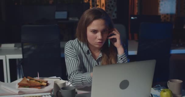 Vista frontal da jovem mulher confusa falando no telefone celular e olhando para a tela do laptop. Menina gerente de escritório sentado na mesa e gestos durante a conversa telefônica no escritório da noite . — Vídeo de Stock