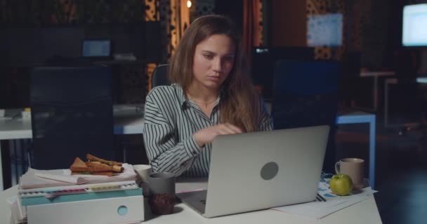 Втомлена жінка має біль і торкається шиї, працюючи пізно на посаді. Дівчина-менеджер офісу використовує свій ноутбук, сидячи за столом на порожньому робочому місці. Концепція перевтоми . — стокове відео