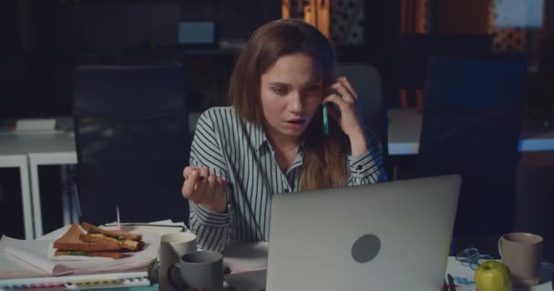 Kadın ofis müdürü dizüstü bilgisayarla çalışıyor ve üzgün bir suratla akıllı telefondan konuşuyor. Genç bir kadın telefon görüşmesi sırasında kötü haberler duyuyor ve hayal kırıklığına uğramış görünüyor. Aşırı çalışma kavramı. — Stok video