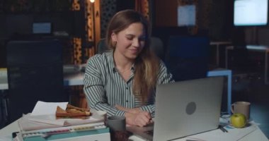 Young woman having video call with boss on her laptop in night office. Angry female employee showing fuck you after video conversation while sitting at her desk in workplace.