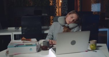 Genç aşırı çalışan bir kadın açık bir dizüstü bilgisayarın önünde otururken uyuyor. Yorgun kız ofis müdürü boş gece ofisinde saatlerce çalışırken boyun yastığıyla uyuyakalıyor..