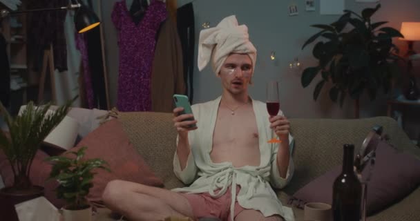 Front view of transgender in rob and towel on head drinking wine and sitting on sofa with smartphone. . Man transgender making jealous face expression while using phone. Home background. — Stock Video