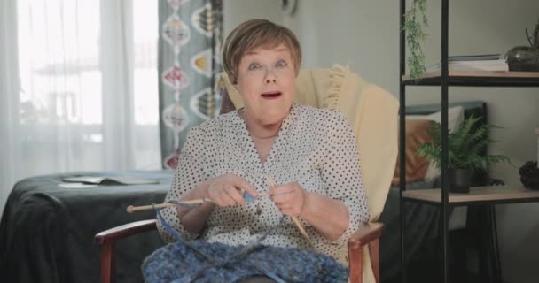 Oude vrolijke dame zittend op stoel en breiend in haar appartement. Vooraanzicht van een oudere vrouw die verrast kijkt terwijl ze breinaalden vasthoudt en glimlacht naar de camera.. — Stockvideo