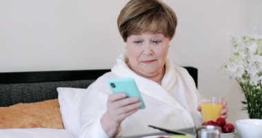Modern akıllı telefon kullanan ve yatakta kahvaltı ederken gülümseyen olgun, güzel bir kadın. Evde soyulan ve elinde bir bardak meyve suyu tutan neşeli yaşlı kadın yatakta şaşkın bir suratla oturuyor..