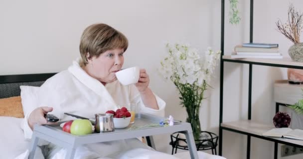 Belle vieille femme qui boit du café pendant le petit déjeuner au lit. Souriant dame âgée à la maison voler regarder la télévision et rire tout en étant assis avec trey plein de nourriture sur ses jambes . — Video