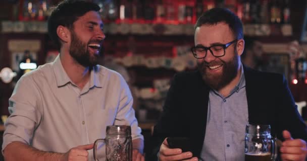 Hombres alegres usando trajes formales en la noche bebiendo cerveza de barril en el mostrador. Berded chico en gafas mostrando foto divertida en el teléfono inteligente a su amigo y riendo mientras se sienta en el pub . — Vídeo de stock