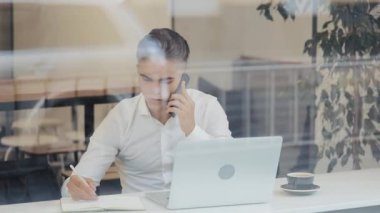 İş sırasında camsı kafede dizüstü bakarak onun Not defteri bilgileri yazma smartphone ile genç iş adamı. Ağır çekim, öğle yemeği zamanı