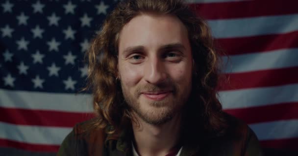 Nahaufnahme eines fröhlichen jungen patriotischen Typen mit Ohrringen, der in die Kamera blickt. Porträt eines bärtigen Millennials, der glücklich und lächelnd aussieht, während er mit amerikanischer Flagge im Hintergrund posiert. — Stockvideo