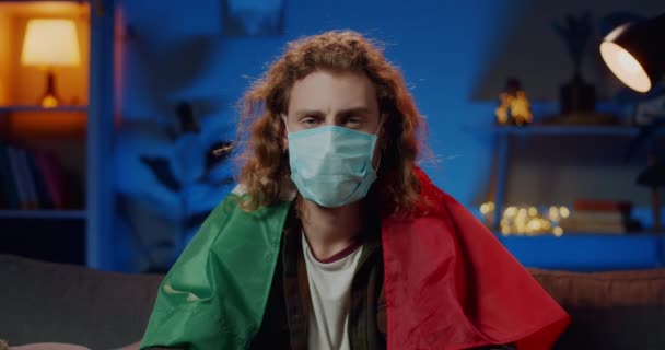 Millenial man in protective mask on face with italian flag on shoulders looking to camera. Portrait of guy staying at home on quarantine. Concept of safety life, coronavirus, virus protection. — Stock Video