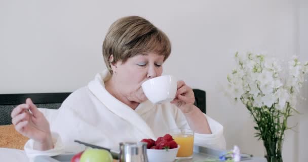 ベッドで朝食をとりながらモーニングコーヒーを楽しむ美しいお年寄りの女性。彼女の足の上に食べ物でいっぱい木に座っている間、ロブの老婦人はカップを保持し、飲みます. — ストック動画