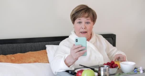 スマートフォンを使いながら、目を信じられないほどのニュースを持つスタイリッシュな高齢女性。朝食を食べながら携帯電話の画面を見ながら驚くべき顔をしている老婦人. — ストック動画